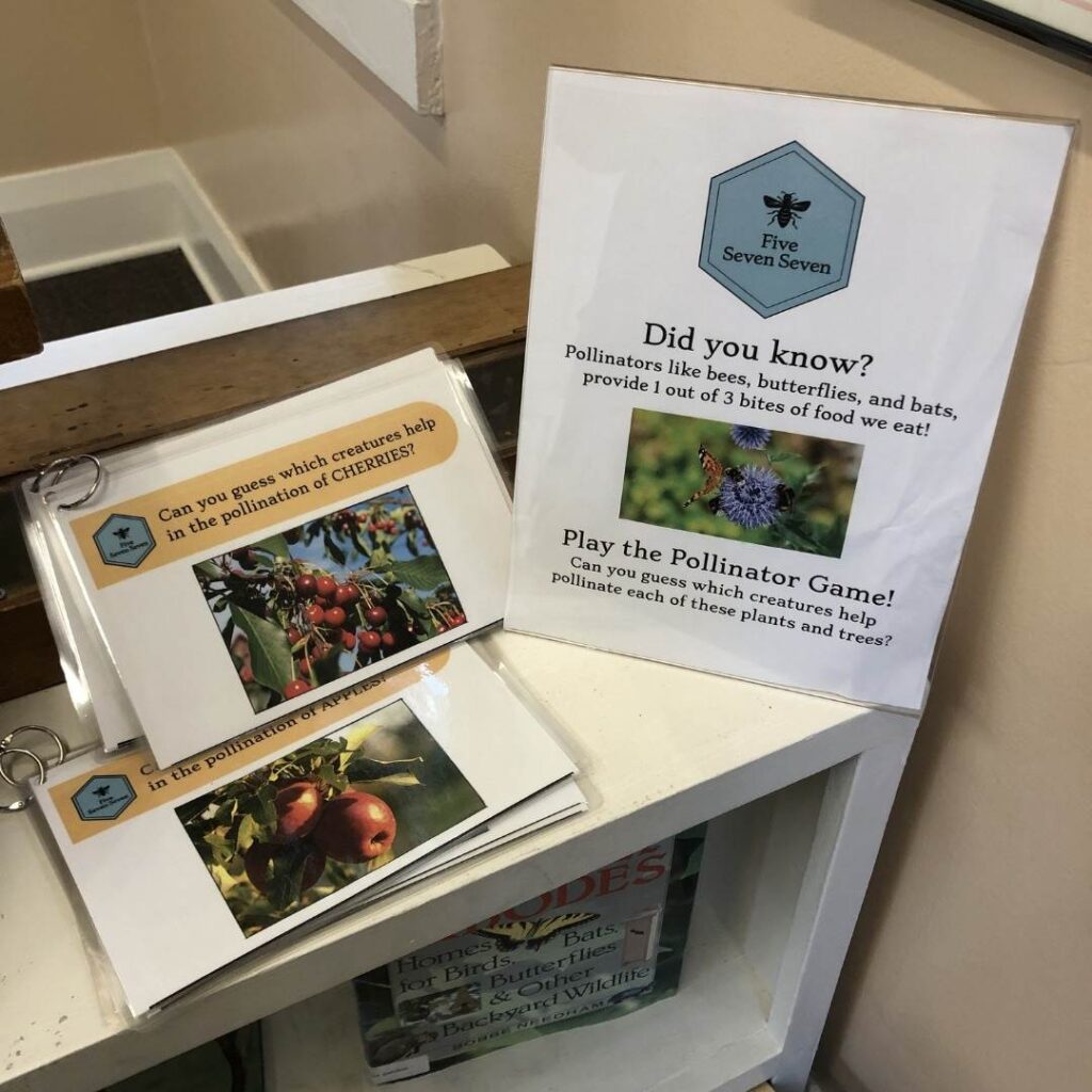 A display table showing a laminated sign that reads “Did you know? Pollinators like bees, butterflies, and bats provide 1 out of 3 bites of food we eat!” Next to it are flip books featuring images of cherries and apples, inviting viewers to guess which pollinators help each crop.