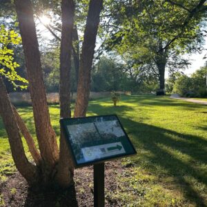 StoryWalk® sign beneath a tree in the sunlit gardens at 577